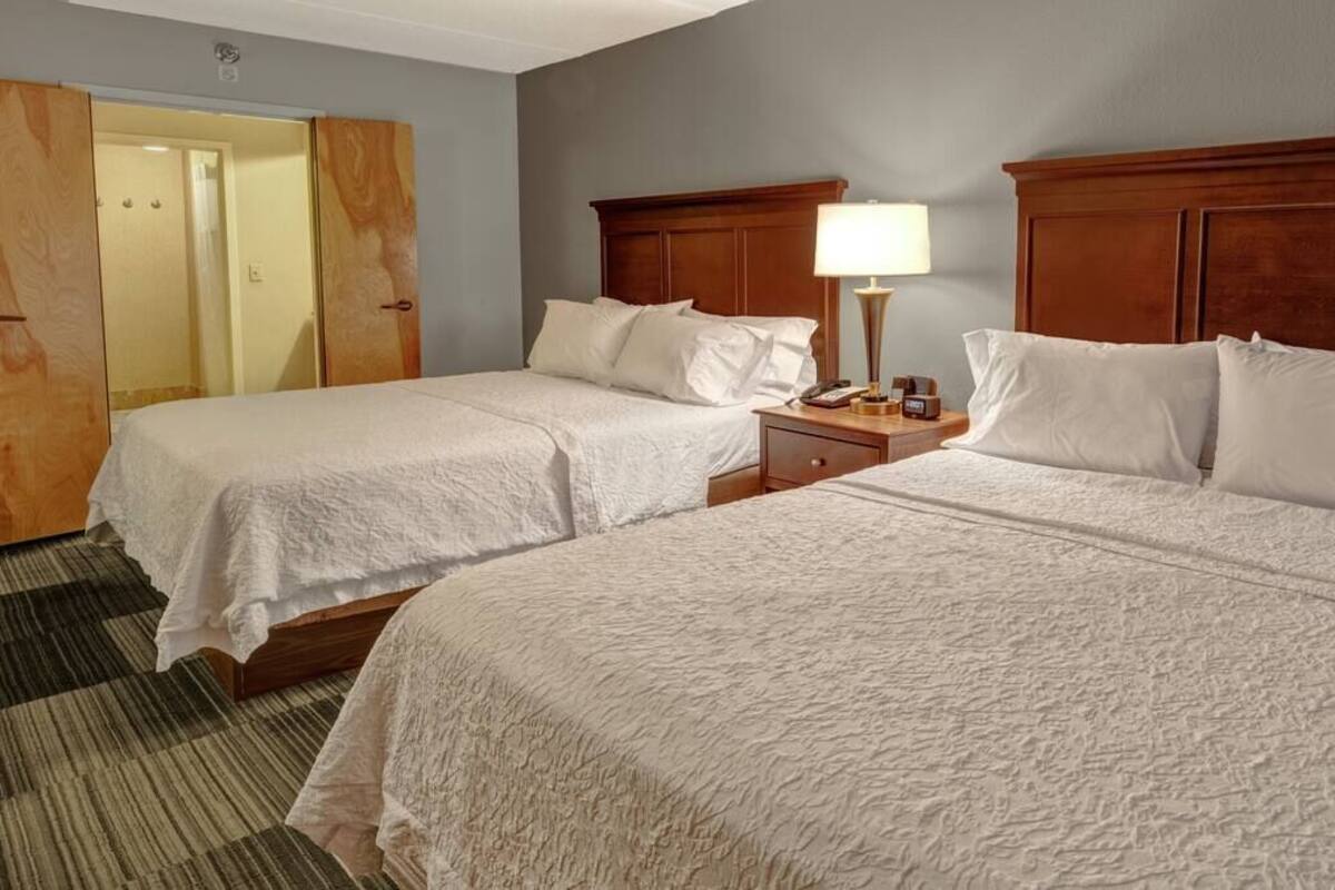 Habitación de hotel con dos camas, cabecero de madera, mesita de noche con lámpara y puerta que da a un baño.