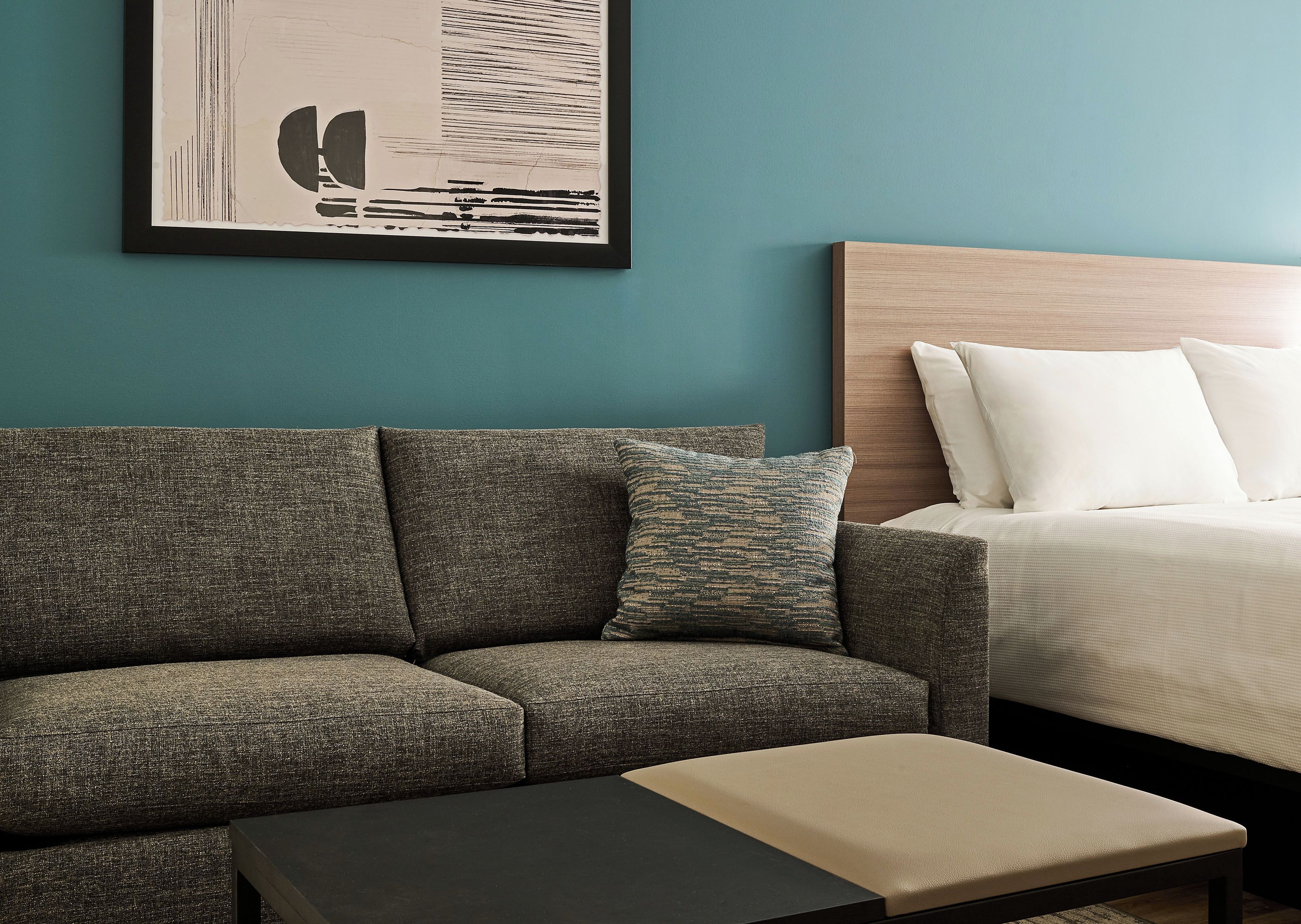 A modern hotel room with a gray sofa, a small table, a bed with white linens, and a framed abstract artwork on the wall.