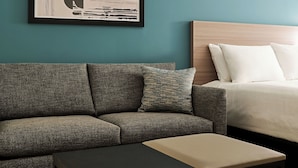 Een moderne hotelkamer met een grijze bank, een kleine tafel, een bed met witte lakens en een abstract kunstwerk in een lijst aan de muur.
