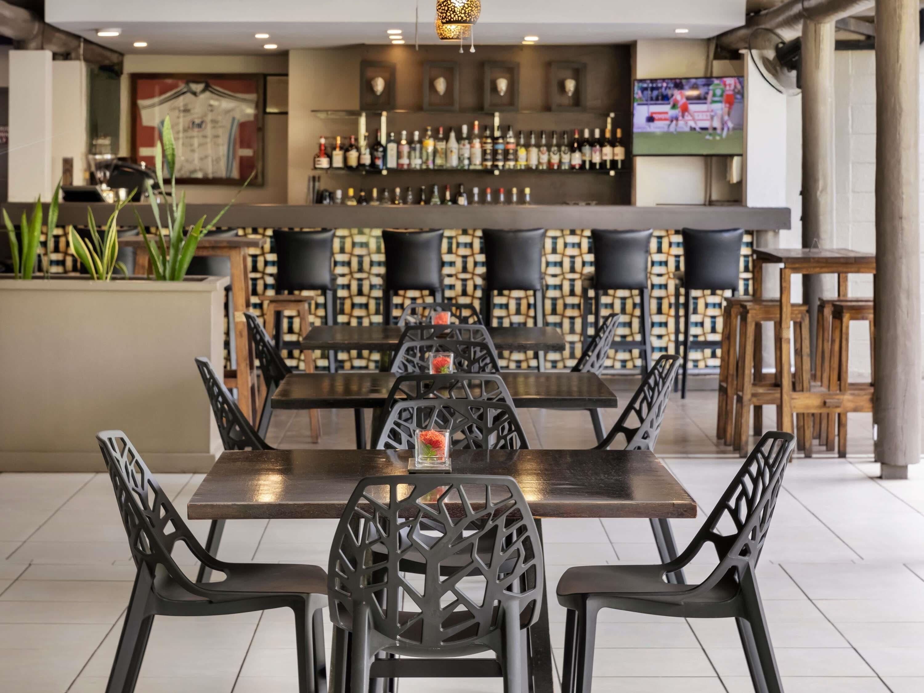 Un restaurante con una mesa larga, sillas negras, una zona de bar con botellas y una pantalla de televisión.