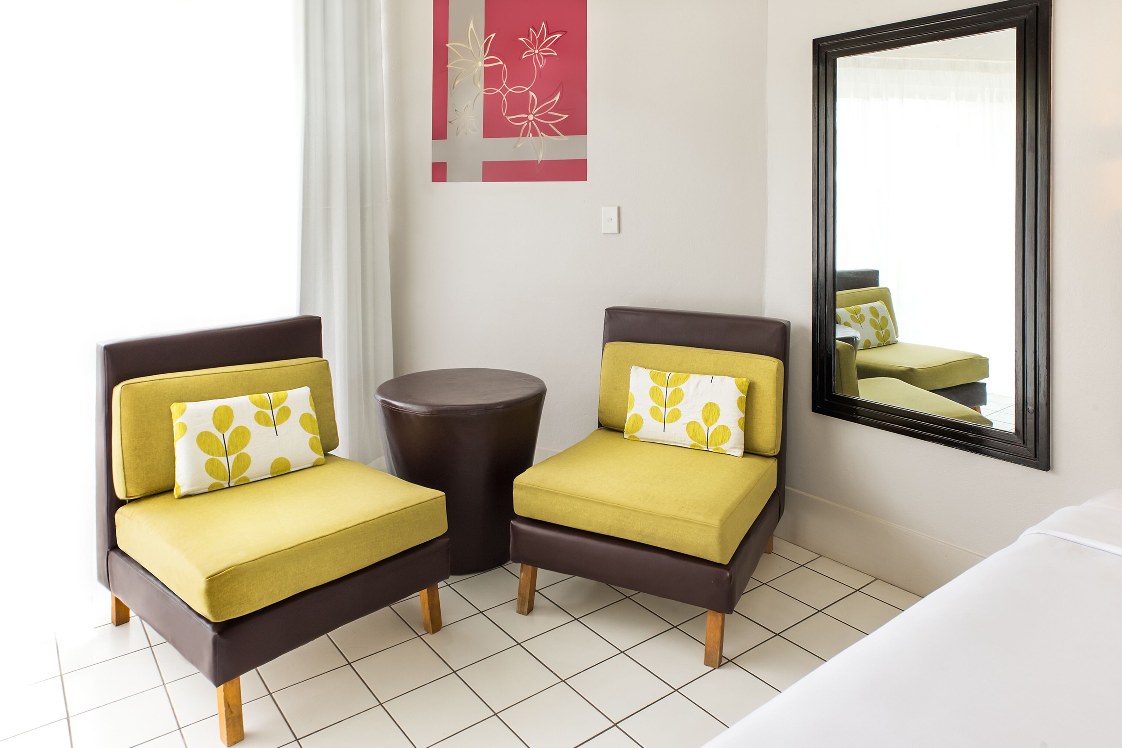 A hotel room with two yellow armchairs, a small round table, a bed, a mirror, and a decorative wall piece.