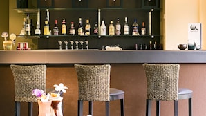 Um espaço de bar com diversas garrafas de bebidas nas prateleiras, cadeiras de vime e uma mesa com um coquetel.