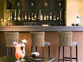 Zona de bar con una variedad de botellas de licor en estantes, sillas de mimbre y una mesa con un cóctel.