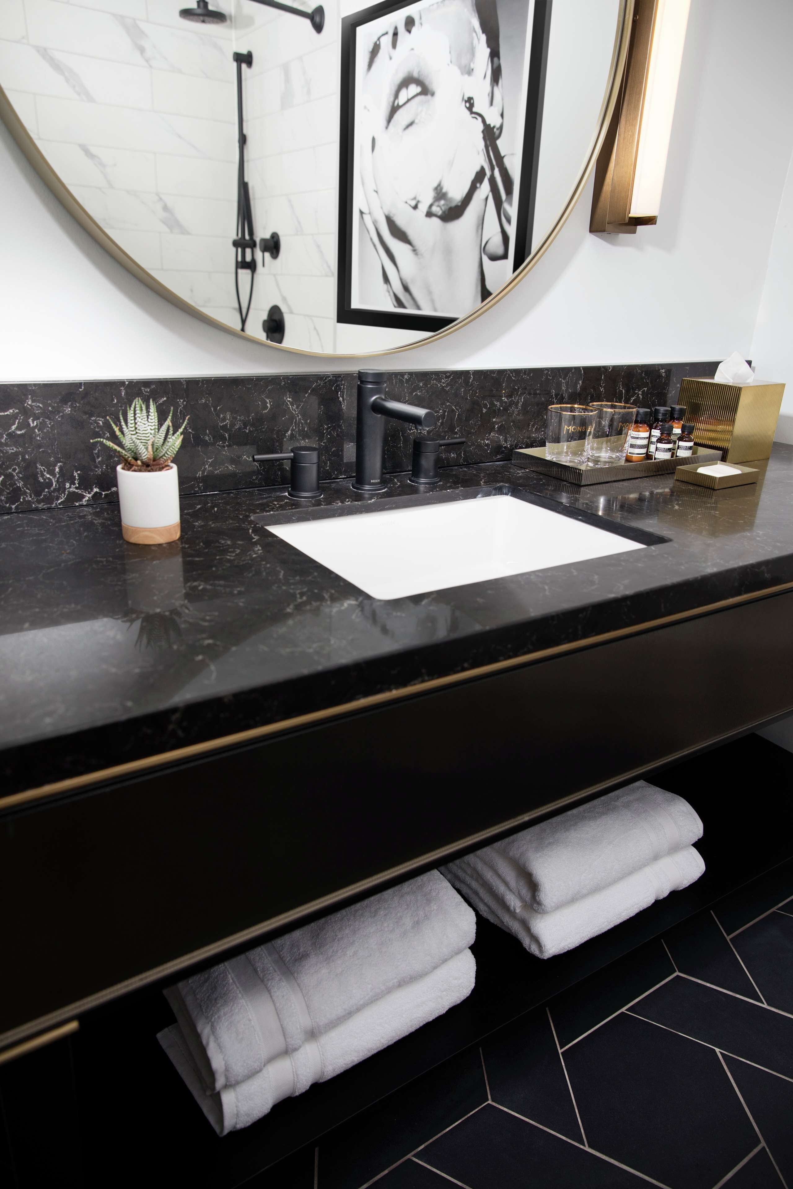 Een moderne badkamer met een zwarte marmeren bladplank, een witte wasbak, een ronde spiegel met gouden lijst, een zwarte kraan en een potplant.