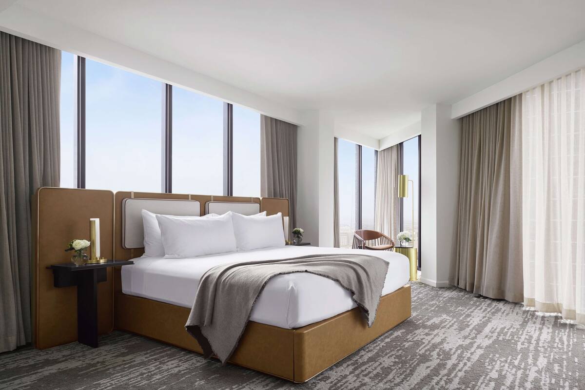 A modern hotel room with a large bed, a nightstand, a chair, and a view of the sky through large windows.