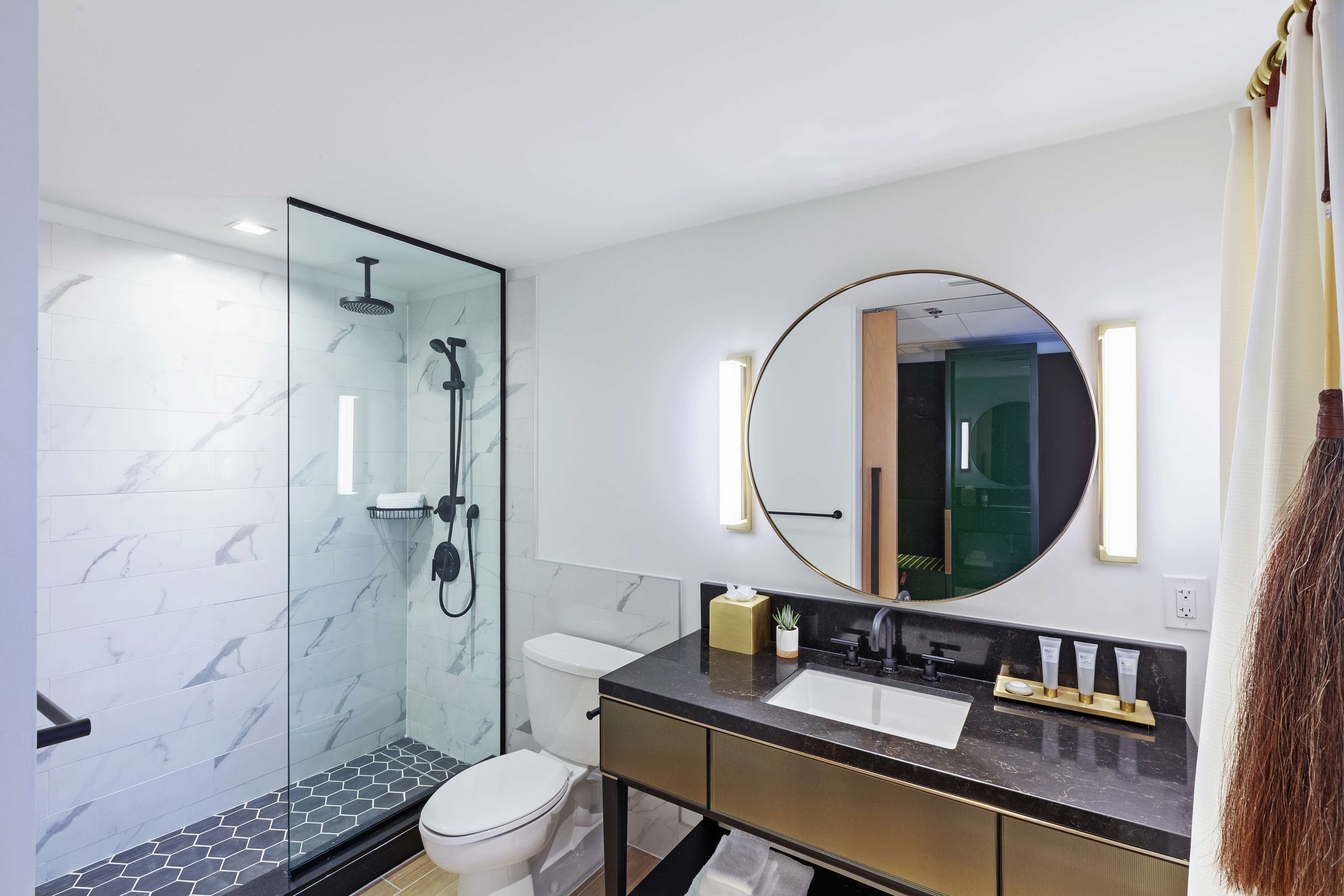 A modern bathroom with a glass-enclosed shower, a toilet, a sink with a gold vanity, and a large round mirror.