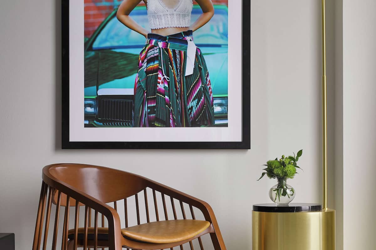 A framed photograph of a person in a white top and colorful skirt, a wooden chair, and a gold floor lamp.