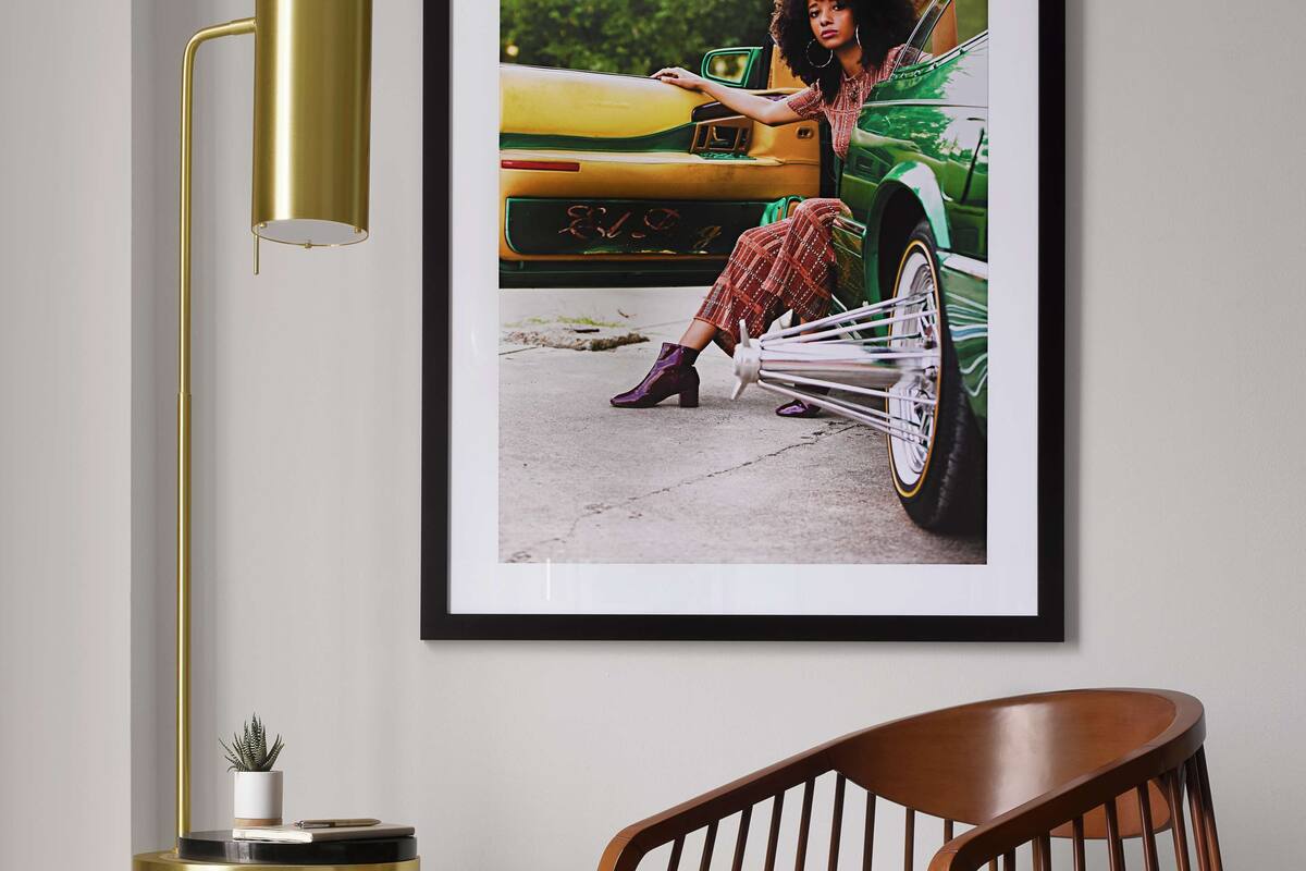 A framed photograph of a vintage car and a woman, a gold floor lamp, and a wooden chair in a room.