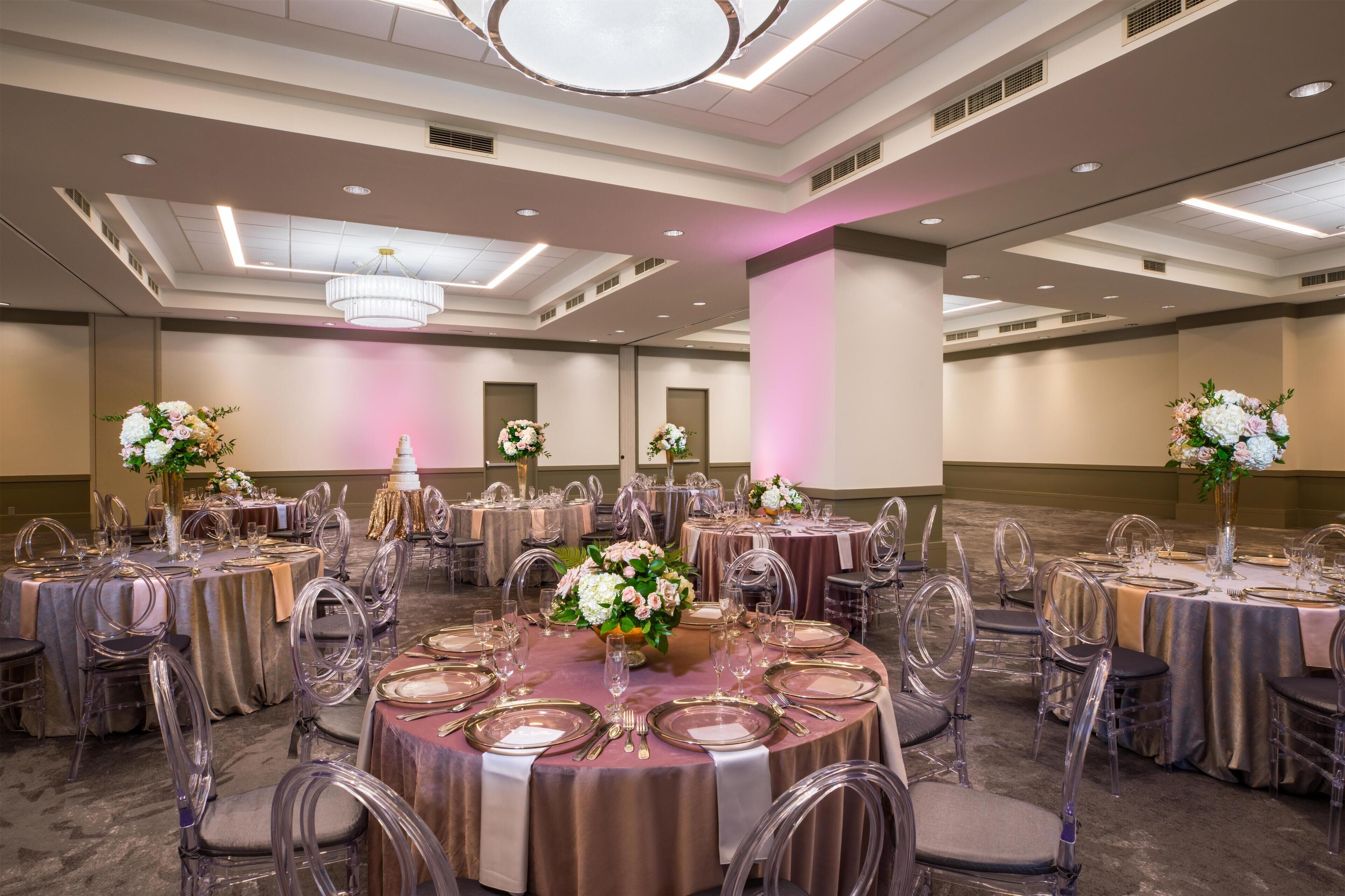 A banquet hall with round tables set for dining, clear chairs, and floral centerpieces.