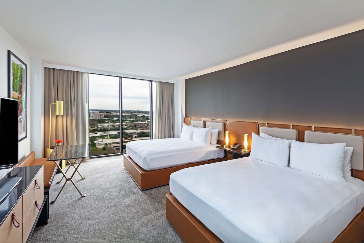 A hotel room with two beds, a TV, a desk, and a large window offering a city view.