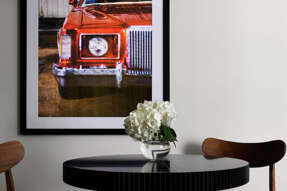 A framed photograph of a vintage car on a wall, a vase with white flowers on a round table, and two wooden chairs.