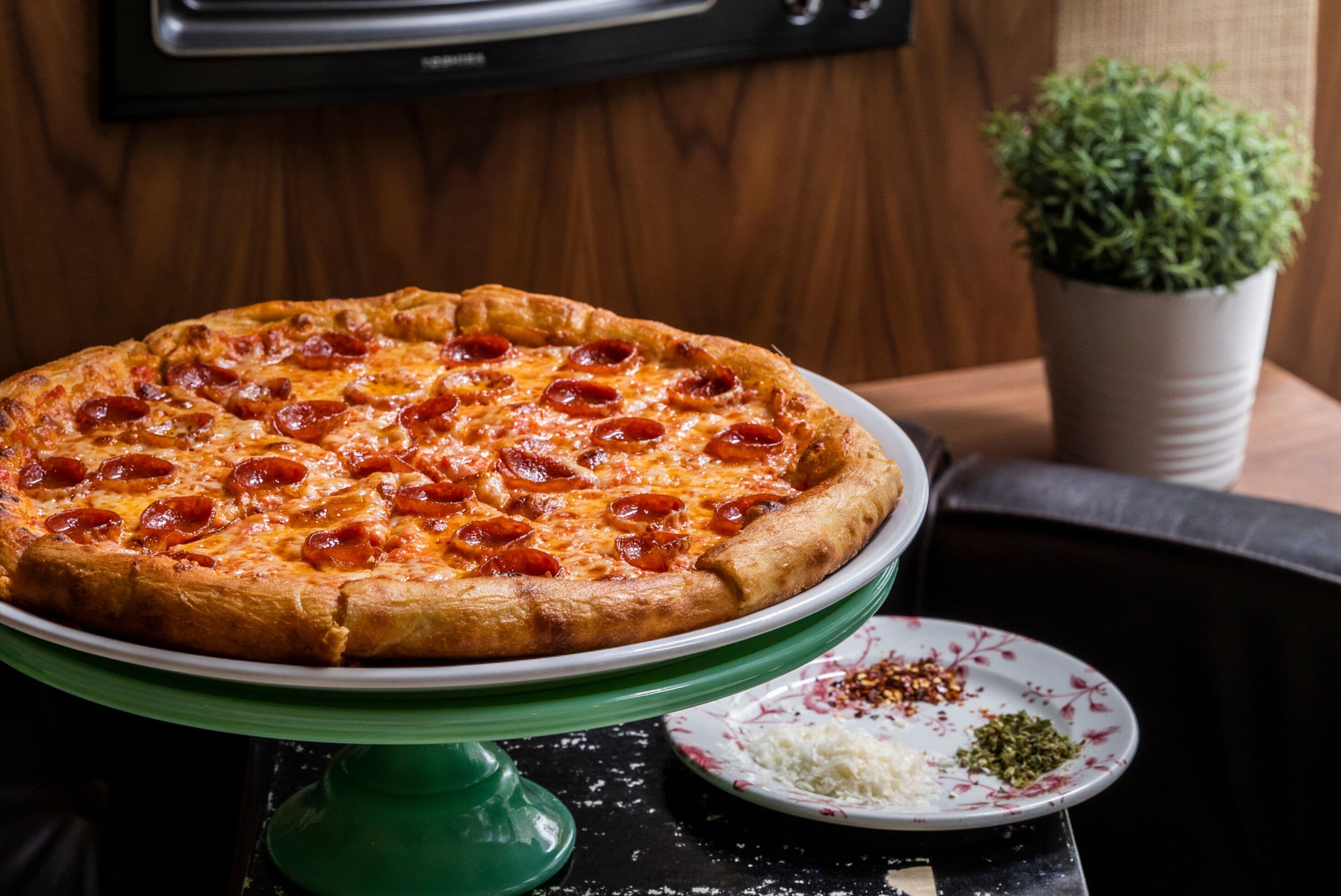 Eine Pizza mit Peperoni auf einem grünen Tisch, daneben ein weißer Teller.