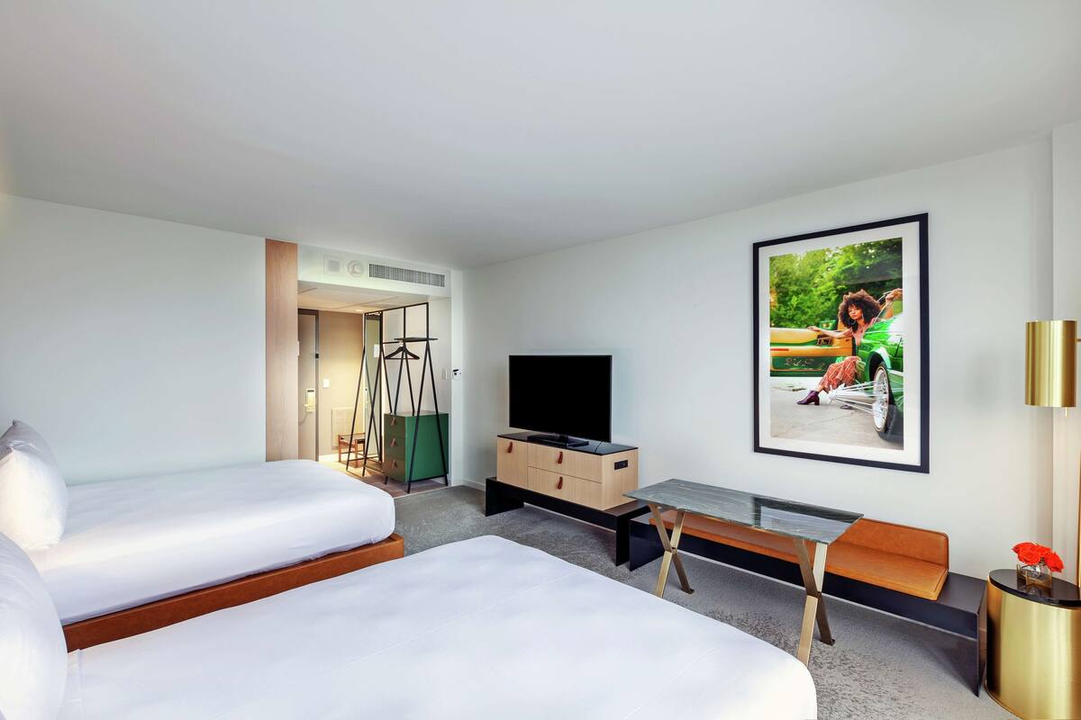 A modern hotel room with two beds, a flat-screen TV, a desk, and a bench.