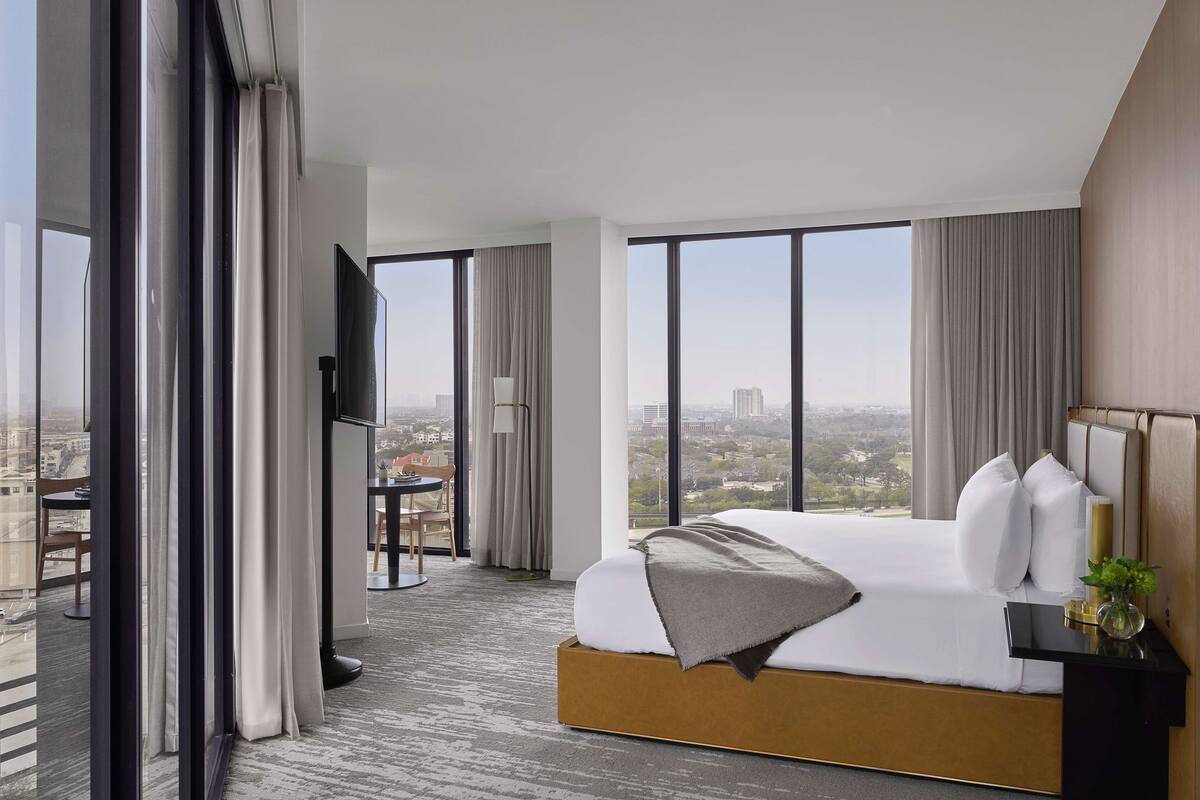 A modern hotel room with a large bed, a TV, a small table, and a view of the city.