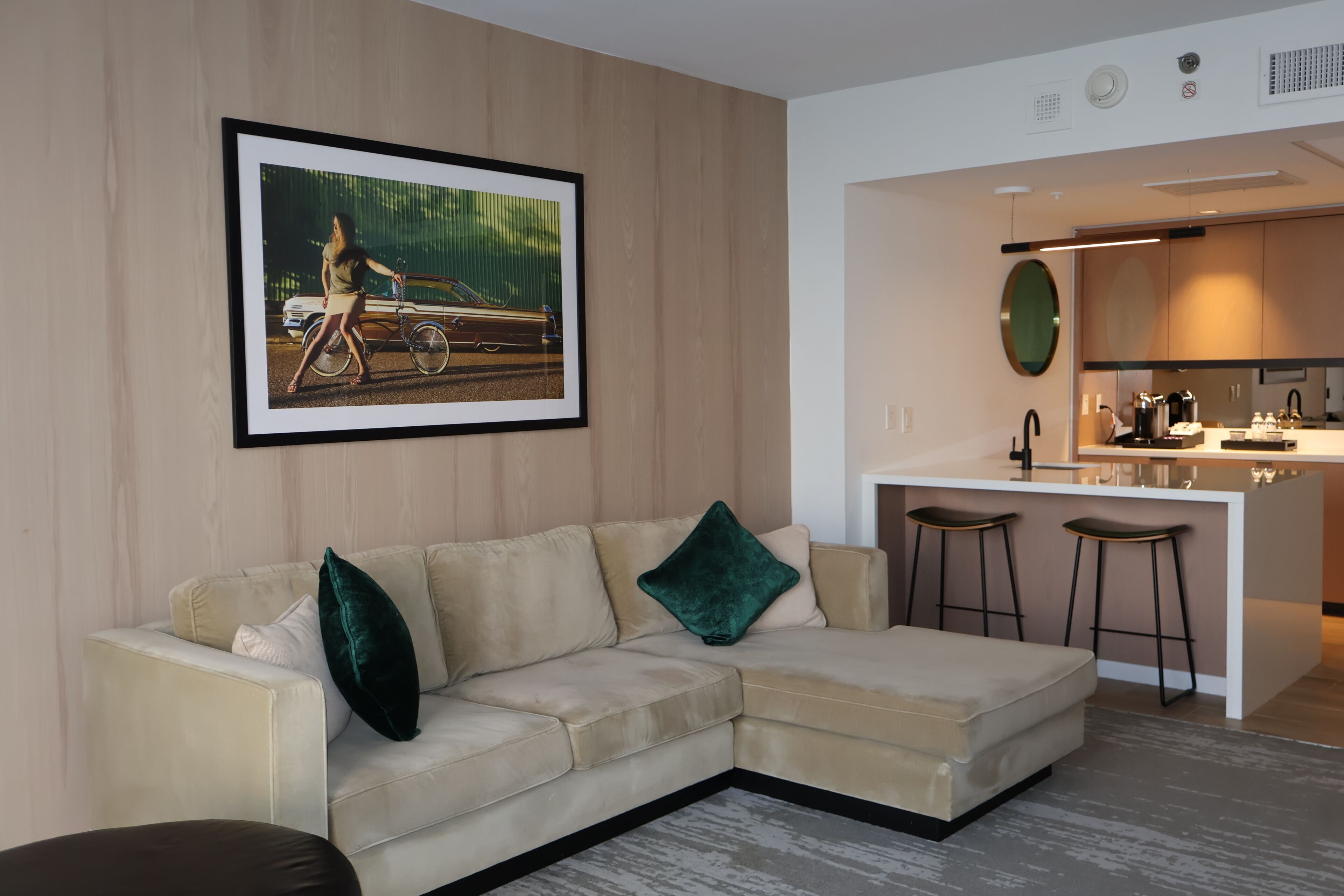 A modern living room with a beige sectional sofa, a framed picture on the wall, and a kitchen area with stools.