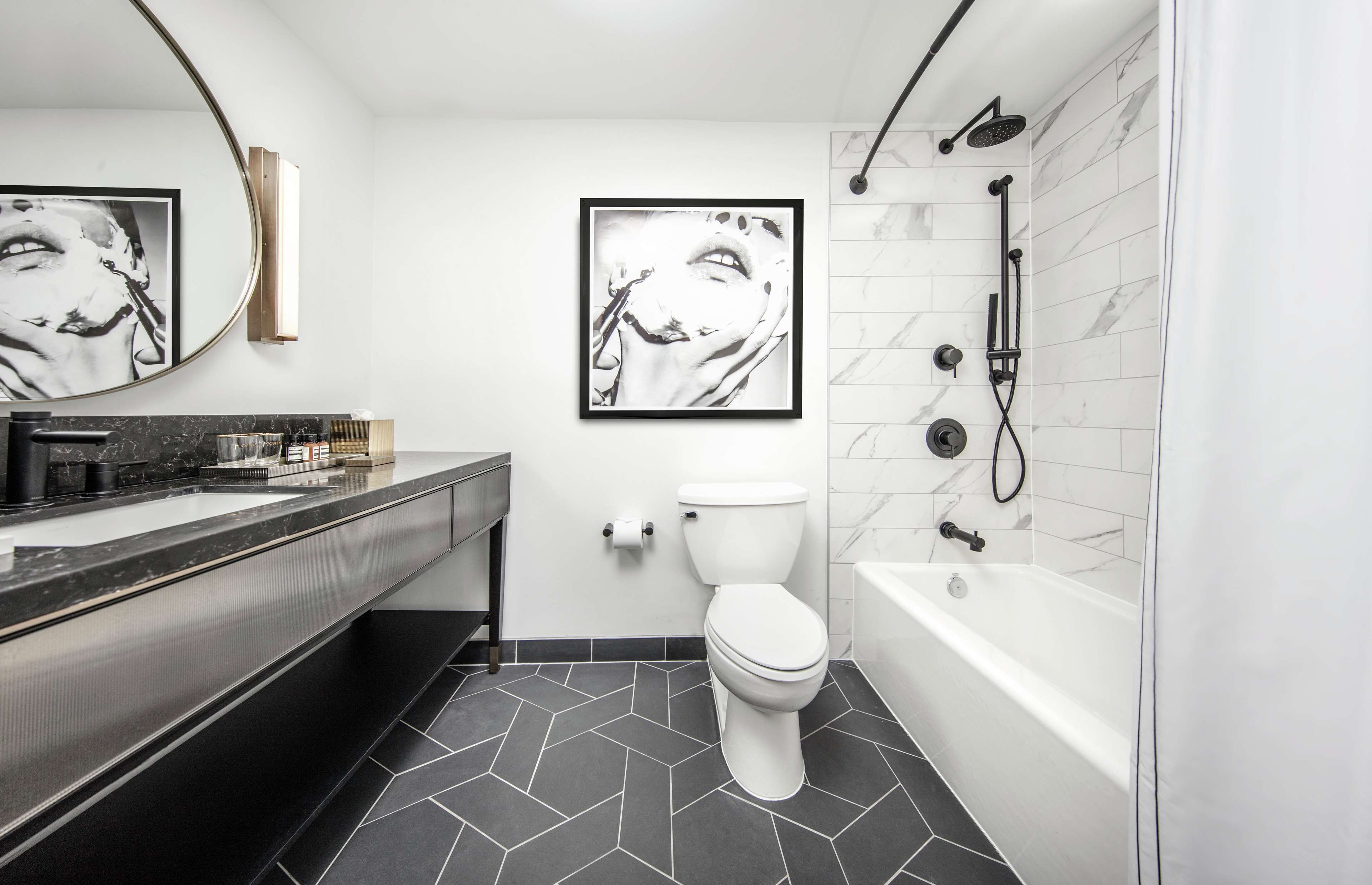 A modern bathroom with a black and white tiled floor, a white toilet, a bathtub, and two framed black and white photographs on the wall.