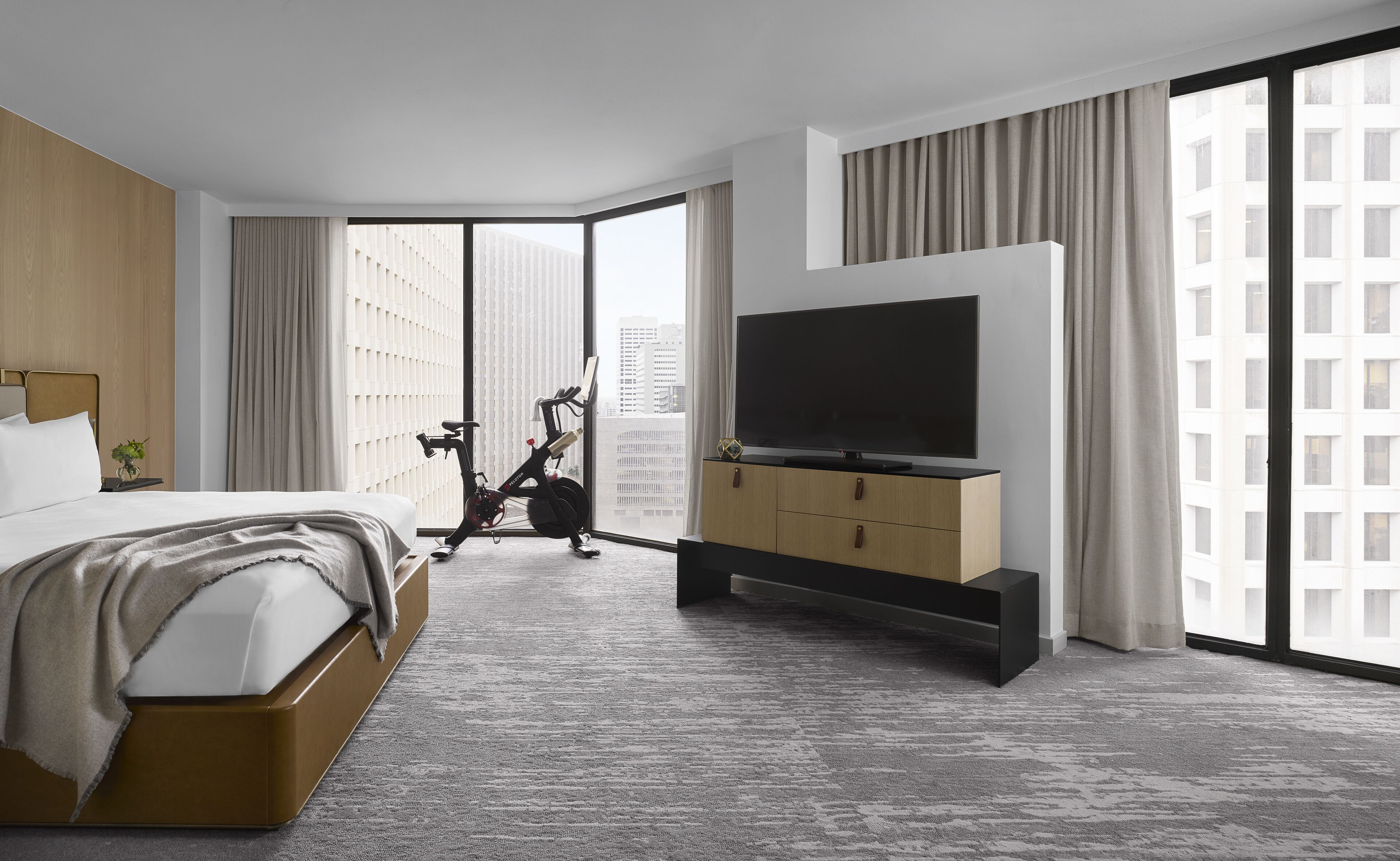 Una habitación de hotel moderna con una cama grande, un televisor sobre un mueble de madera, una bicicleta y vistas a la ciudad a través de amplias ventanas.