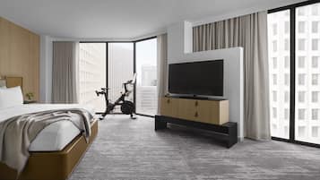 Una habitación de hotel moderna con una cama grande, un televisor sobre un mueble de madera, una bicicleta y vistas a la ciudad a través de amplias ventanas.