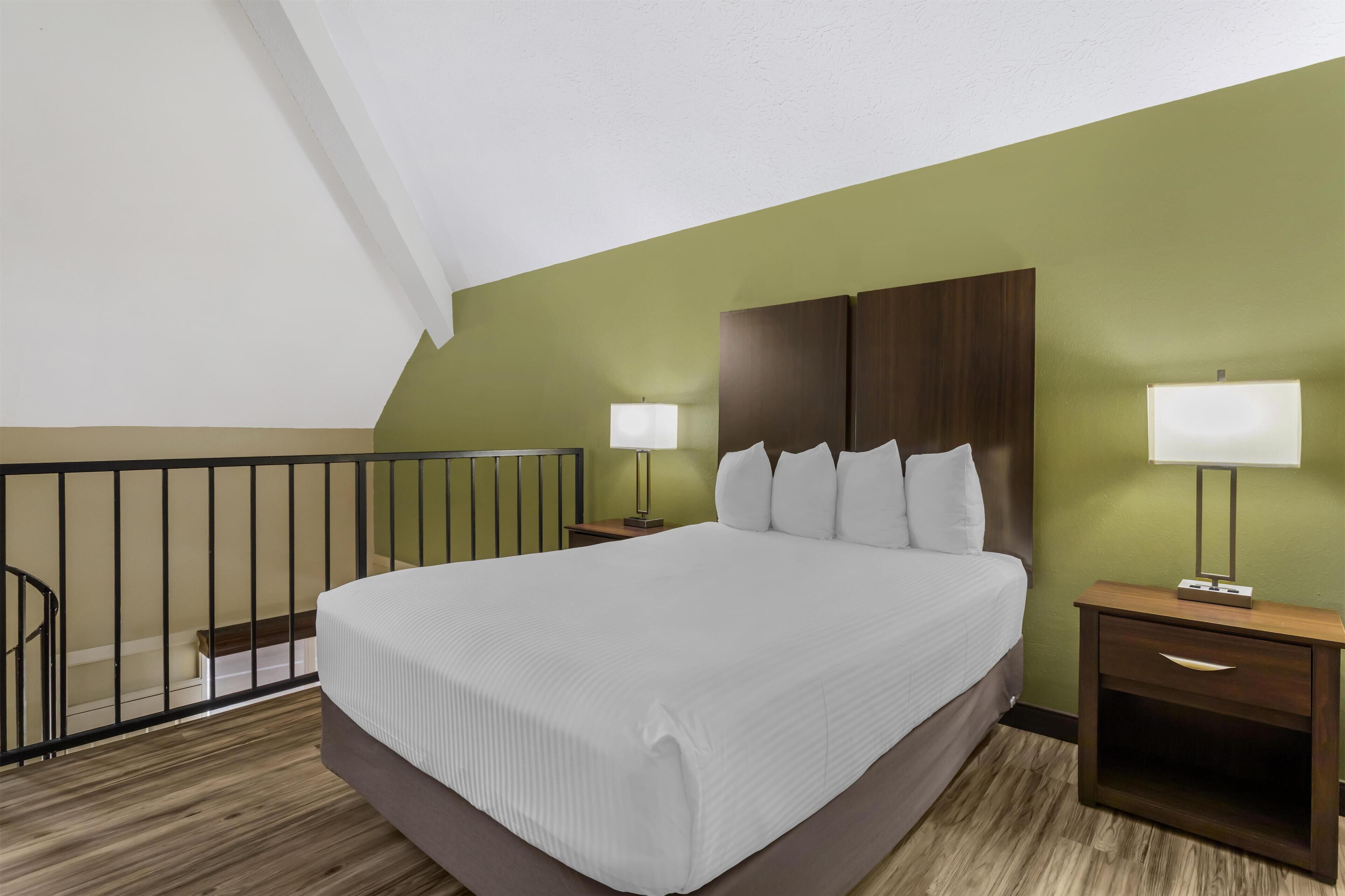 Quarto de hotel com uma cama grande, duas mesas de cabeceira com luminárias, um painel de cabeceira de madeira e uma varanda com guarda-corpo.