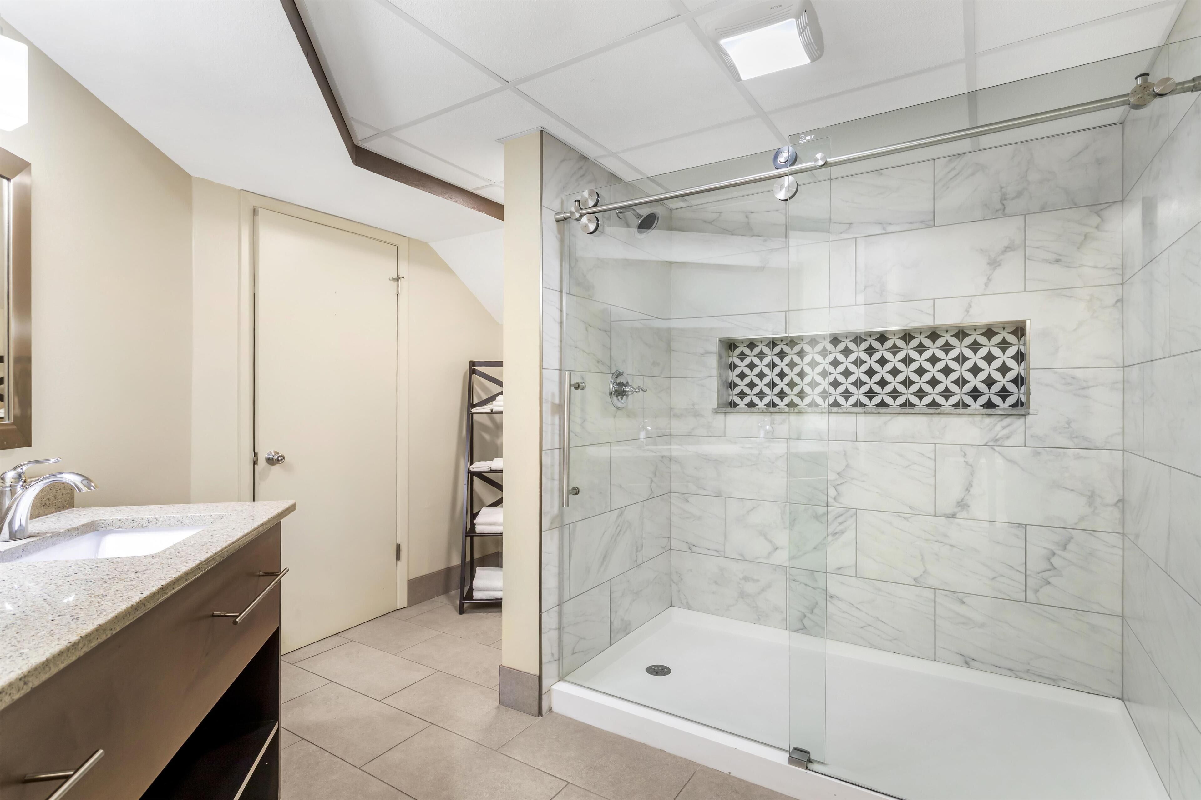 A modern bathroom with a glass-enclosed shower, a marble-tiled wall, a built-in shelf with towels, and a vanity with a sink and cabinet.