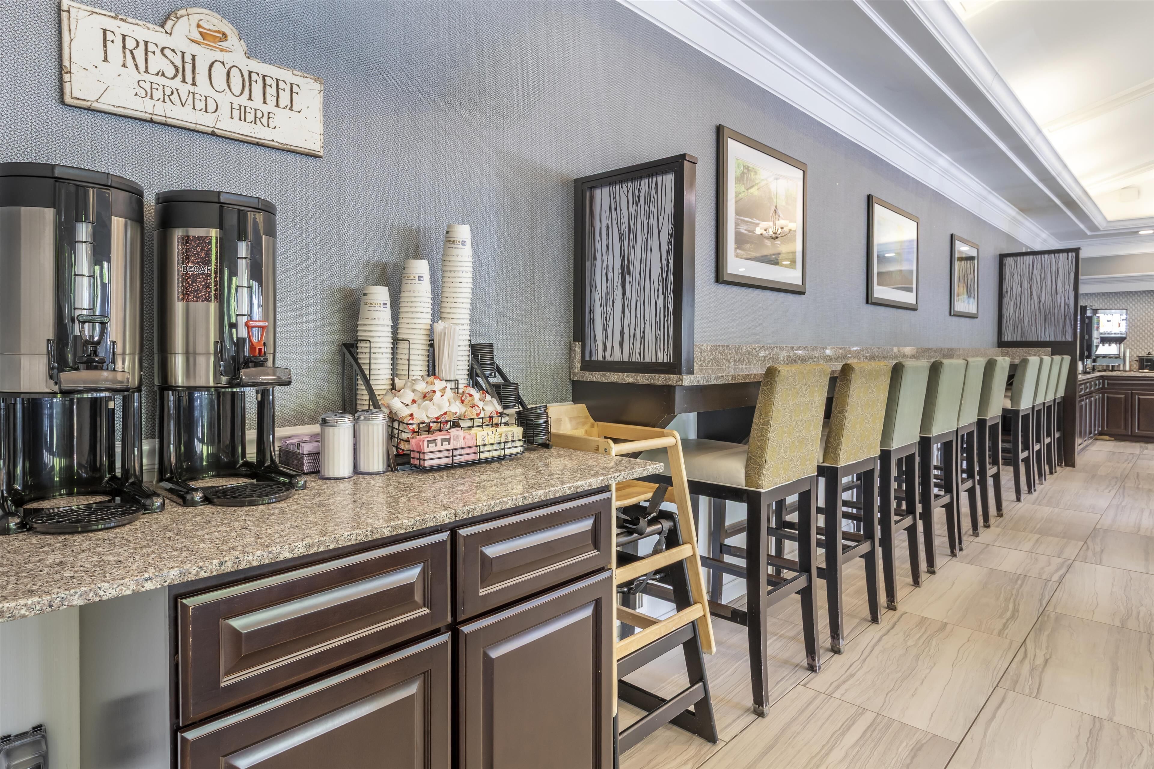 A coffee station with multiple coffee dispensers, a sign reading "FRESH COFFEE SERVED HERE," and a row of chairs along the wall.