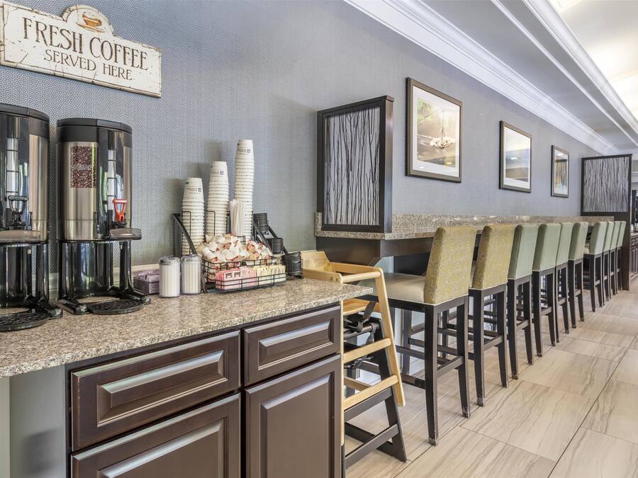 A coffee station with multiple coffee dispensers, a sign reading "FRESH COFFEE SERVED HERE," and a row of chairs along the wall.