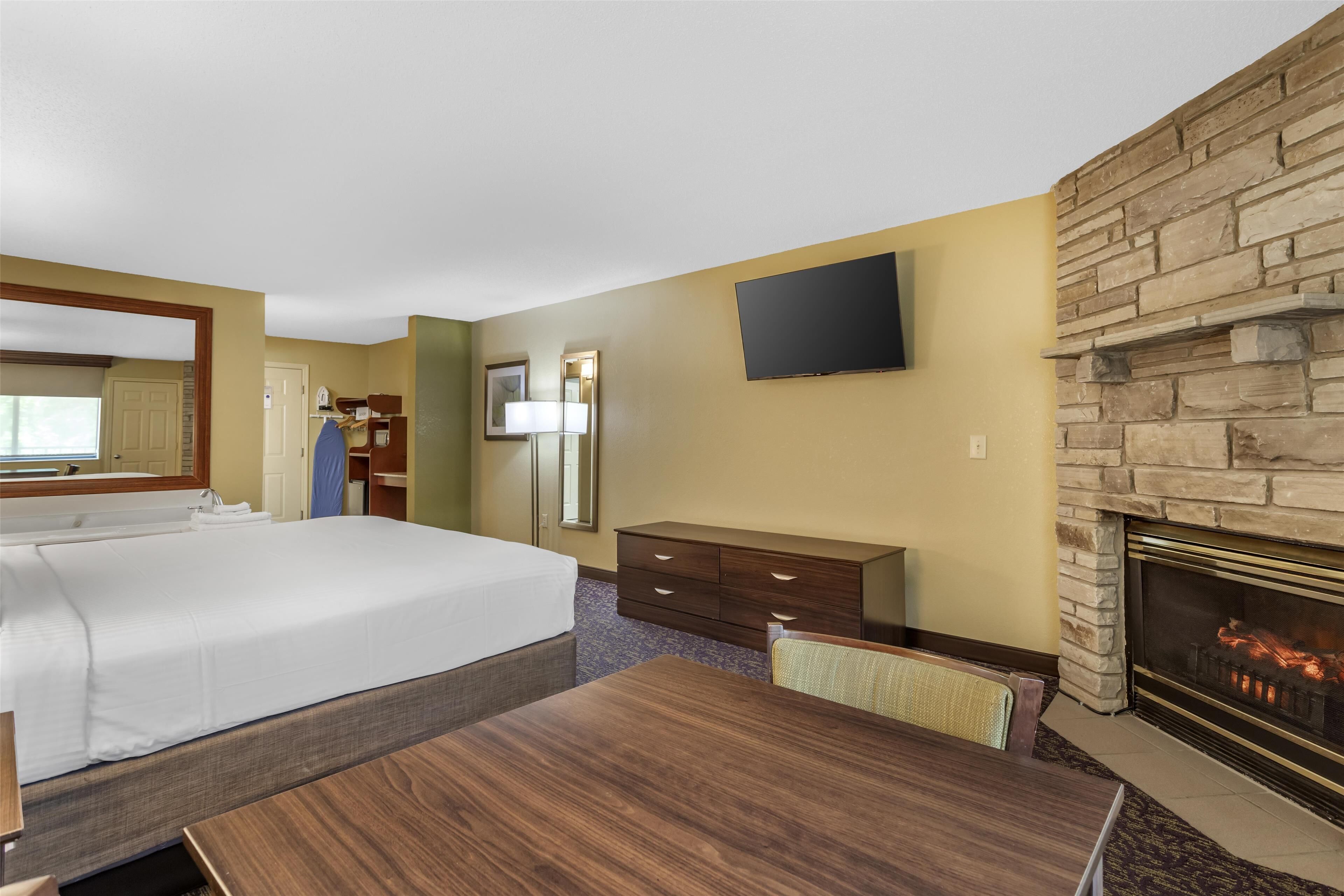 A hotel room with a large bed, a wooden dresser, a flat-screen TV, a stone fireplace, and a mirror.