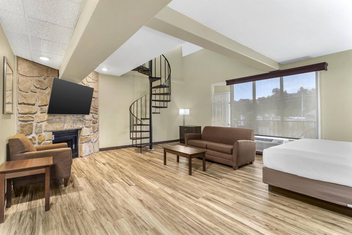 Un salon spacieux avec une cheminée en pierre, un escalier moderne, un grand lit et une vue sur l’extérieur par une fenêtre.