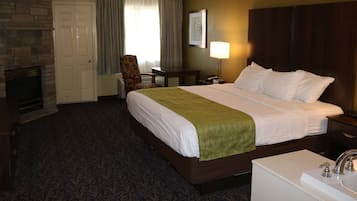 A hotel room with a large bed, a bedside table, a lamp, a chair, a fireplace, and a bathroom with a sink and mirror.