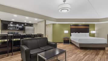 A compact hotel room with a kitchenette, a bed, a sofa, a small table, and a nightstand with lamps.