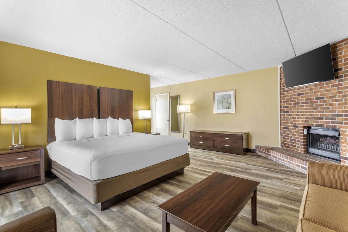 Une chambre d’hôtel avec un grand lit, une tête de lit en bois, un téléviseur à écran plat fixé à un mur en briques, une cheminée et une table basse en bois.