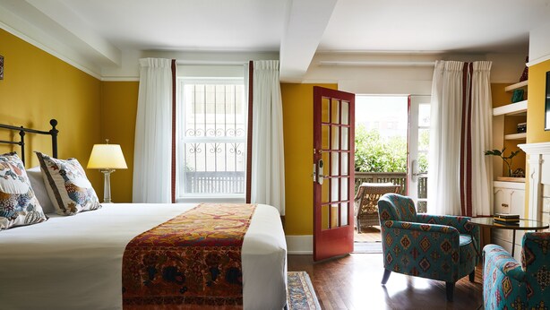 Ein Schlafzimmer mit einem großen Bett, einer Sitzecke mit gemusterten Stühlen und Blick ins Freie durch eine Tür.