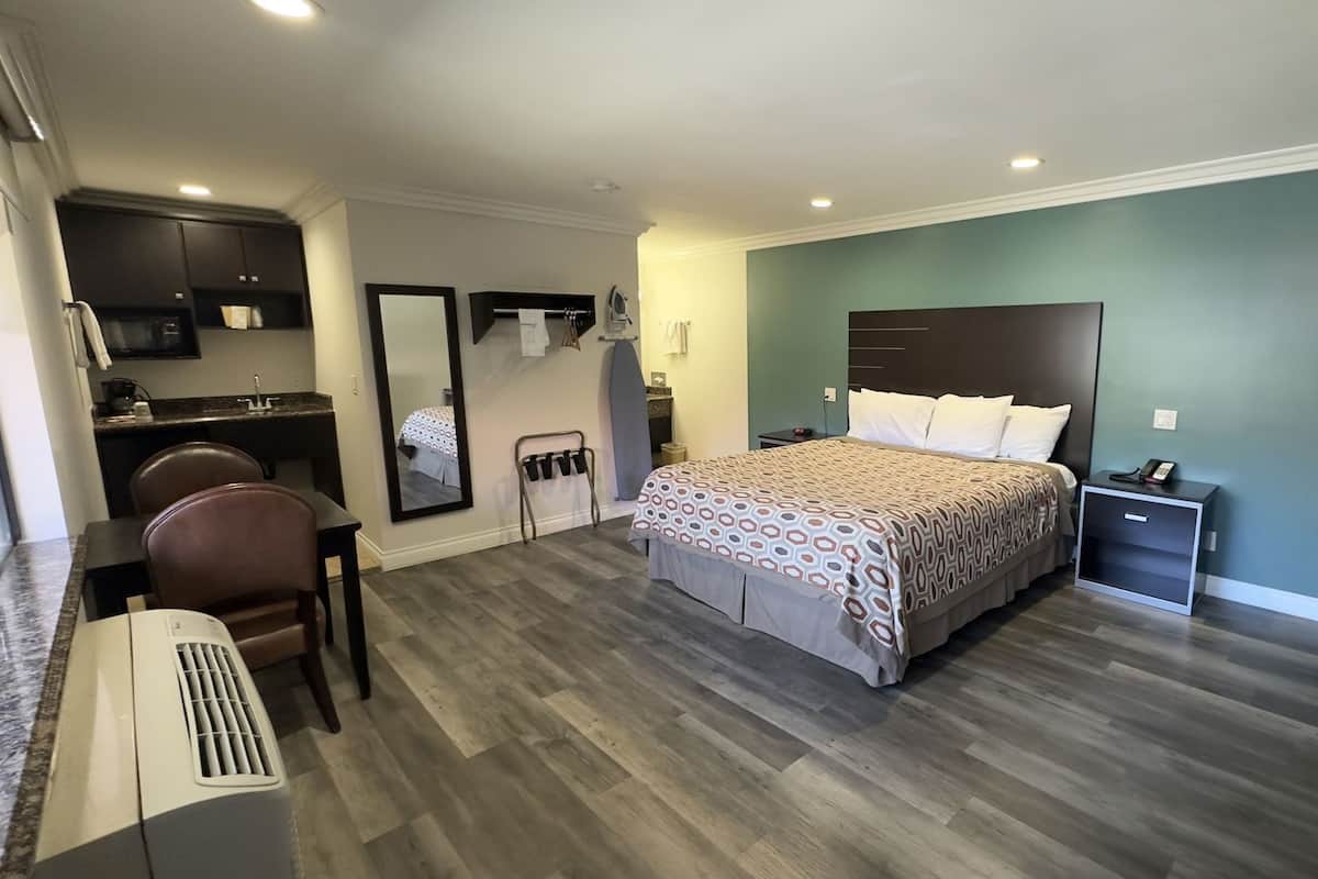 A bedroom with a bed, a desk, a chair, a mirror, and a small kitchenette.