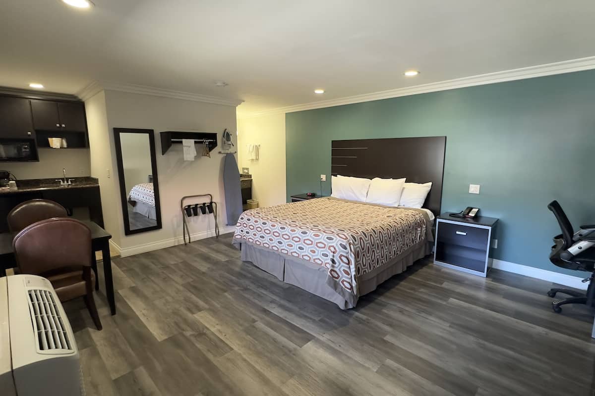 A bedroom with a bed, a desk, a chair, a mirror, a nightstand, and a small kitchenette.
