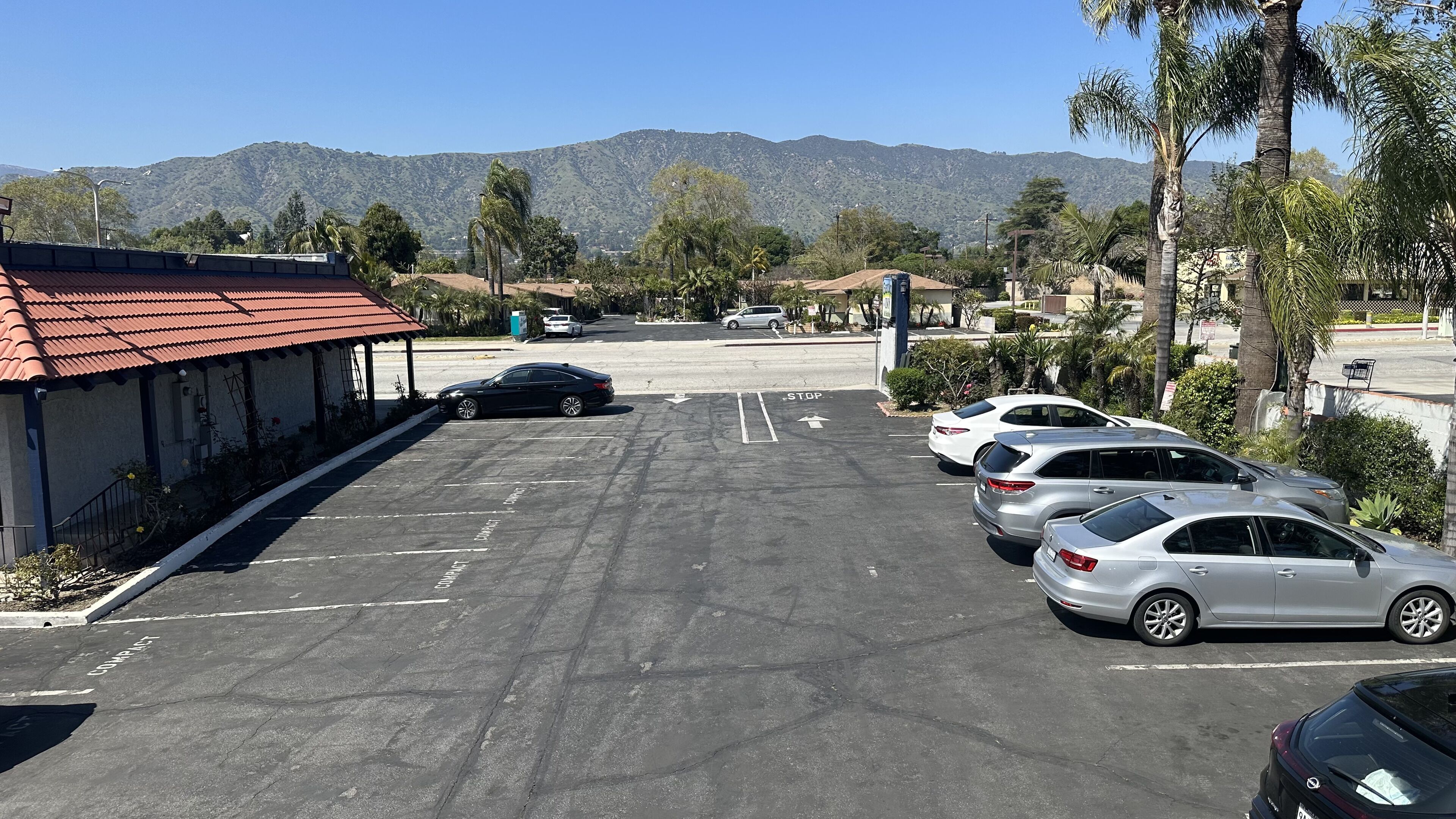 A parking lot with several cars, a building with a red roof, and mountains in the background.