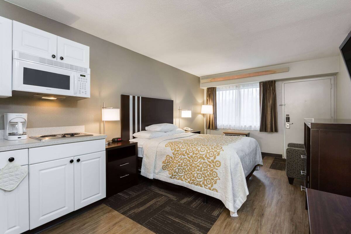 A hotel room with a bed, bedside table, lamp, and a kitchenette with a microwave.
