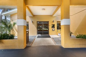 Fachada de edificio de color amarillo, puerta de cristal con la leyenda 'ENTRANCE' y un camino de hormigón que conduce a la puerta.