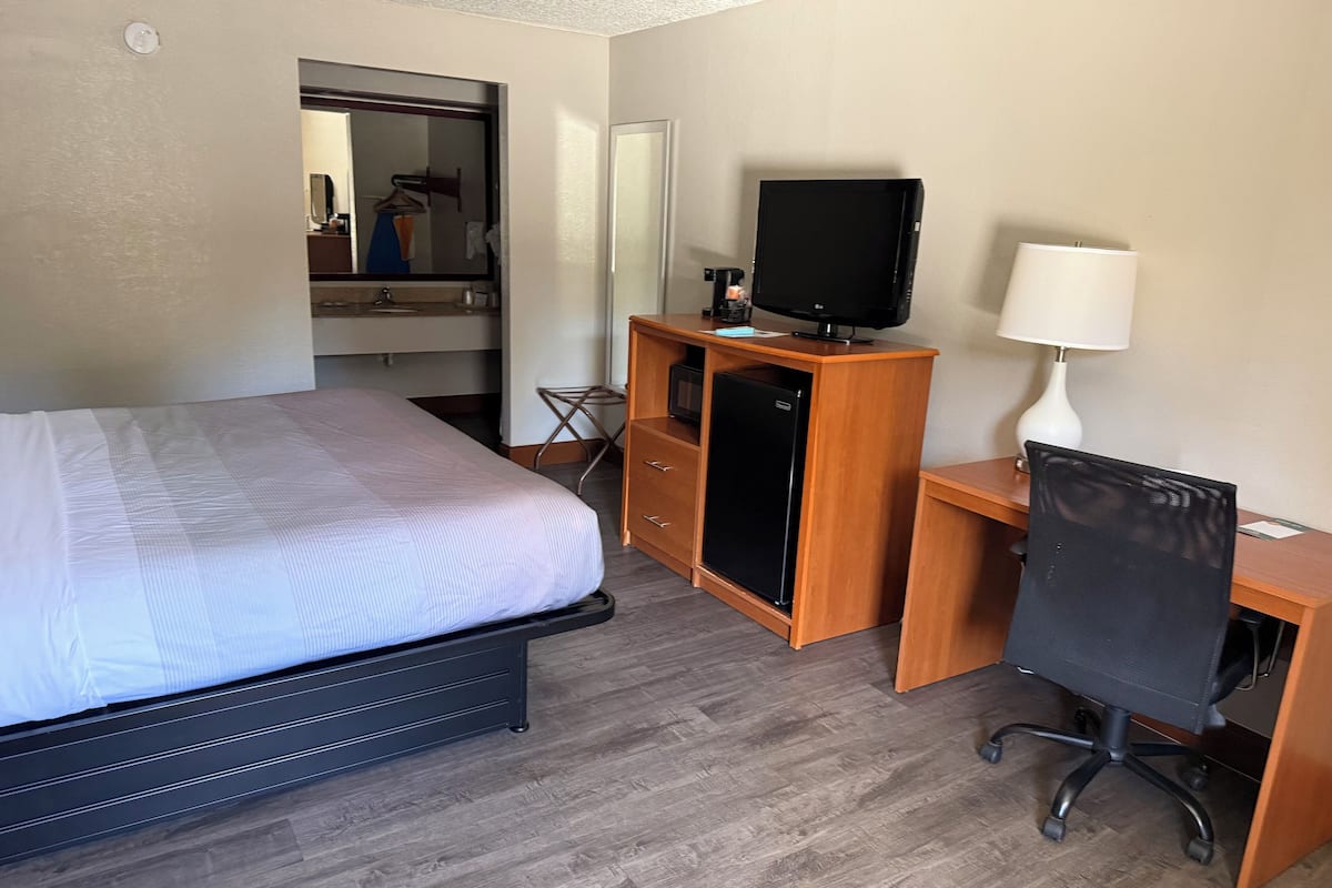A hotel room with a bed, a desk with a computer and a lamp, a small refrigerator, and a mirror.