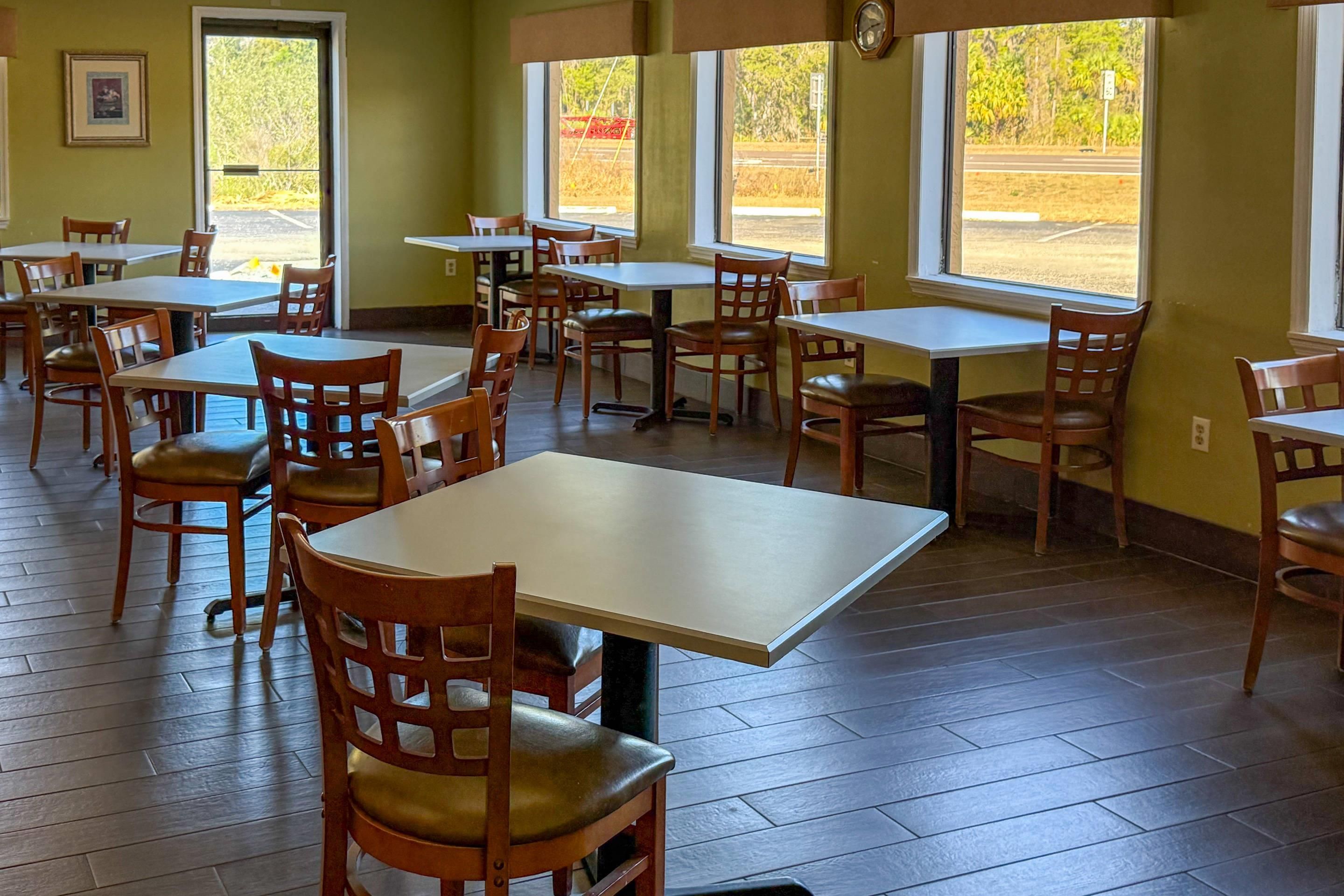 A dining area with multiple tables and chairs, large windows with views of the outdoors, and a clock on the wall.