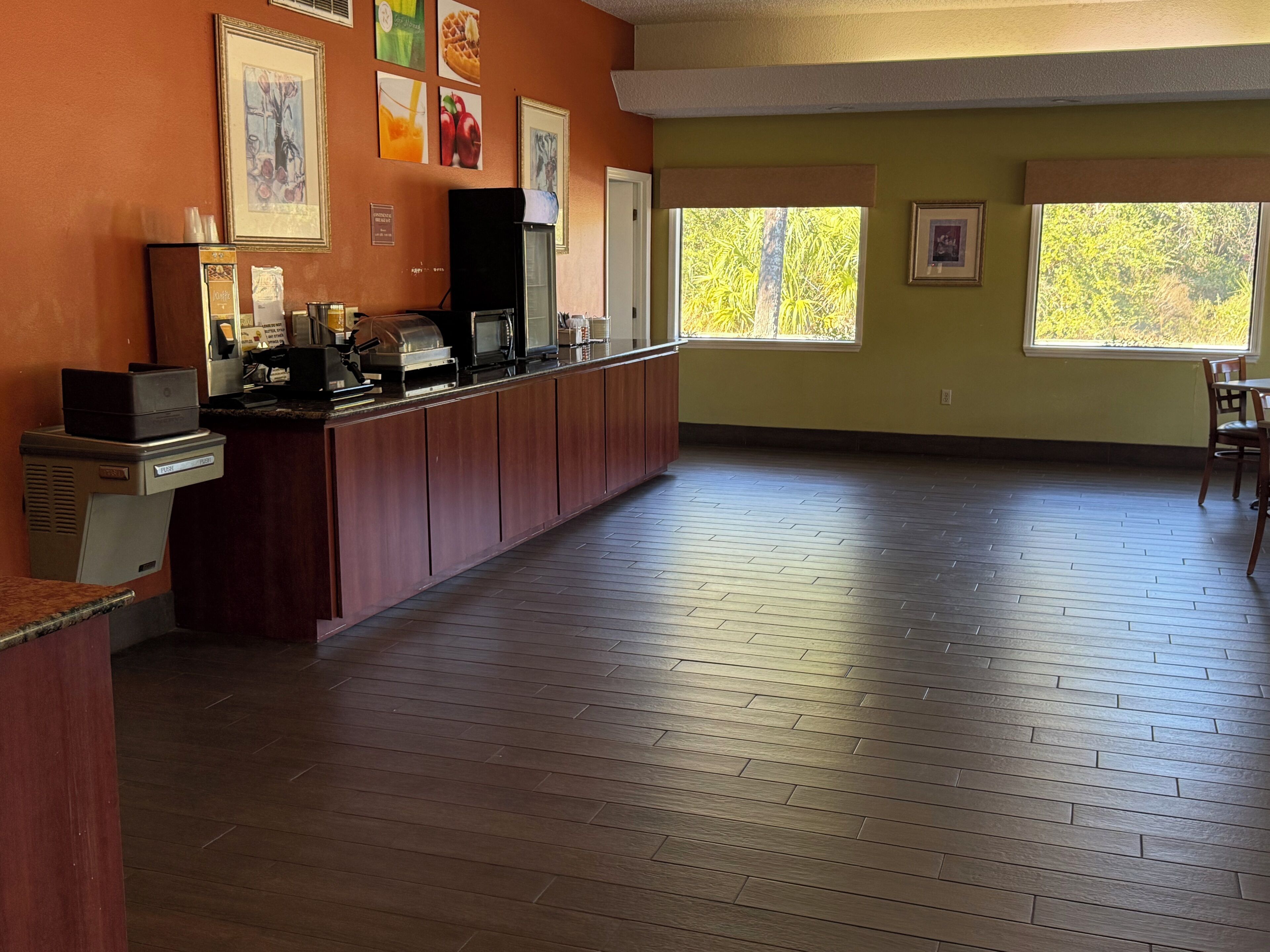 A spacious room with a long wooden counter, a kitchenette, and large windows offering a view of trees.