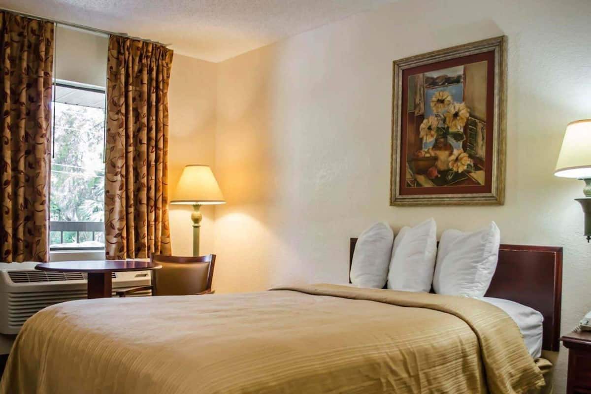 A hotel room with a bed, a desk with a chair, a window with curtains, a painting on the wall, and a lamp on the nightstand.