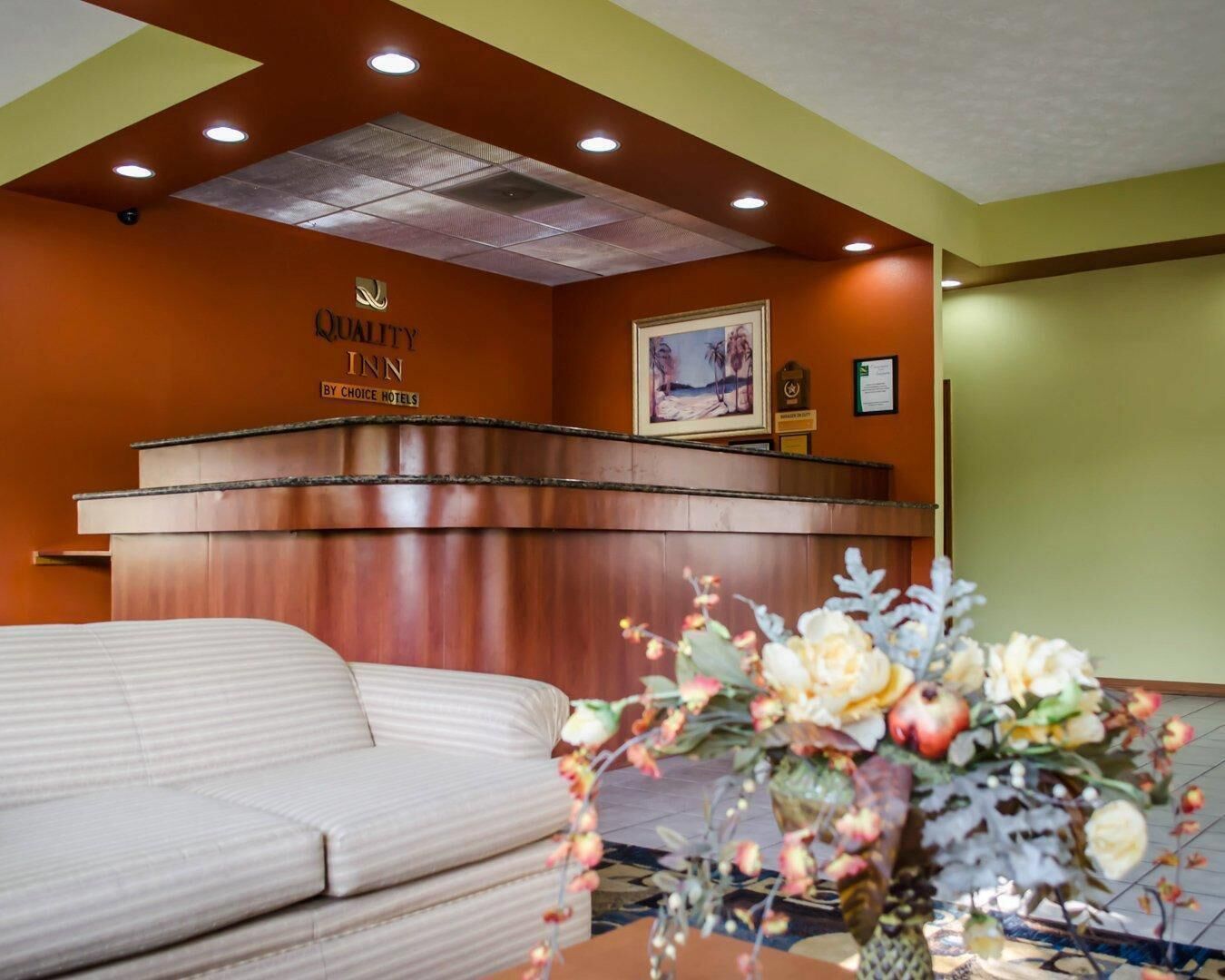 A hotel reception area with a reception desk, a framed picture of a beach scene, and a floral arrangement on a table.