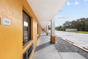 Un pasillo con una pared exterior amarilla, la habitación 122 señalizada y un estacionamiento con grava y una cerca al fondo.