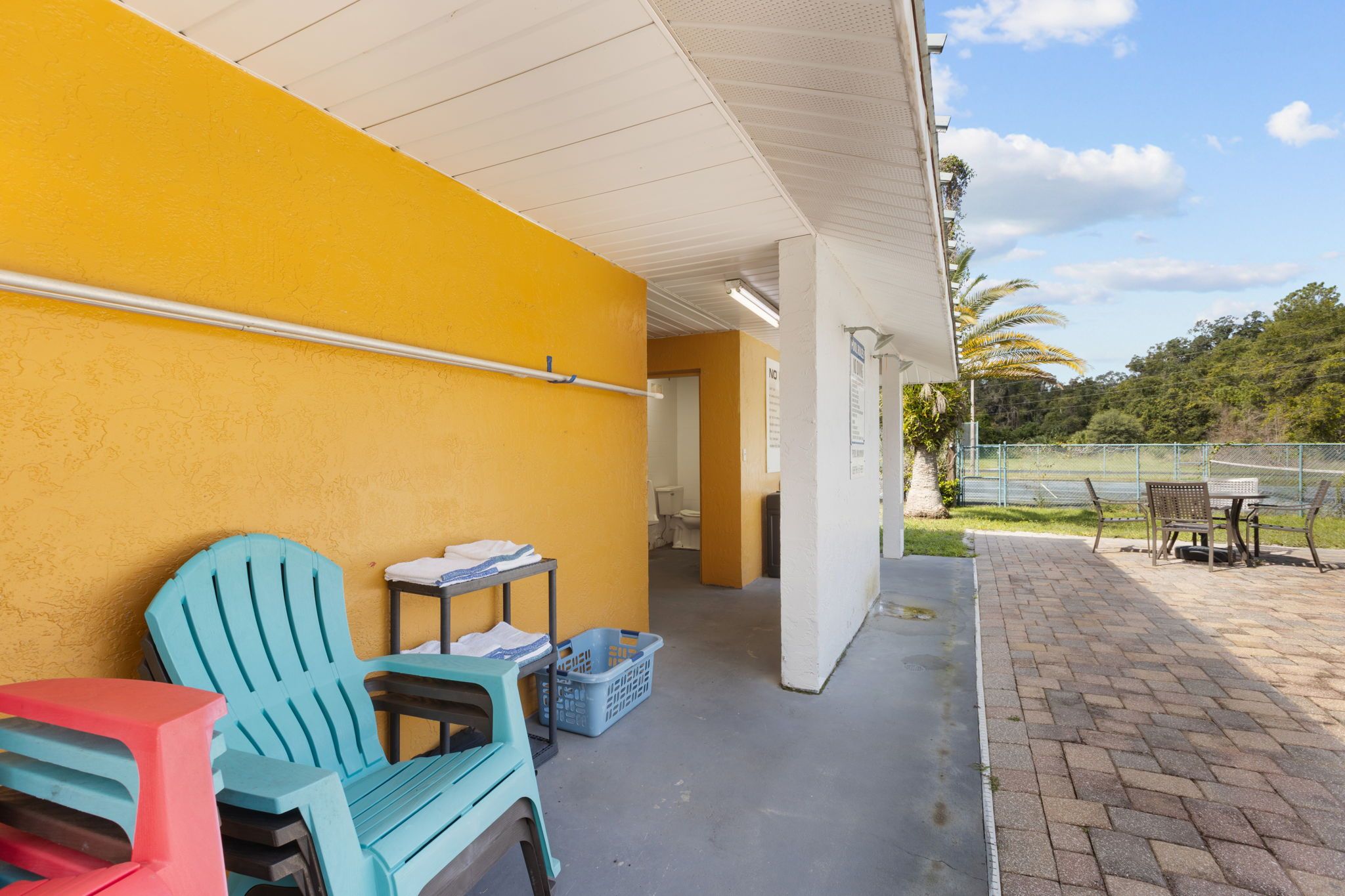 A covered outdoor area with a yellow wall, a blue chair, a table, and a trash bin.