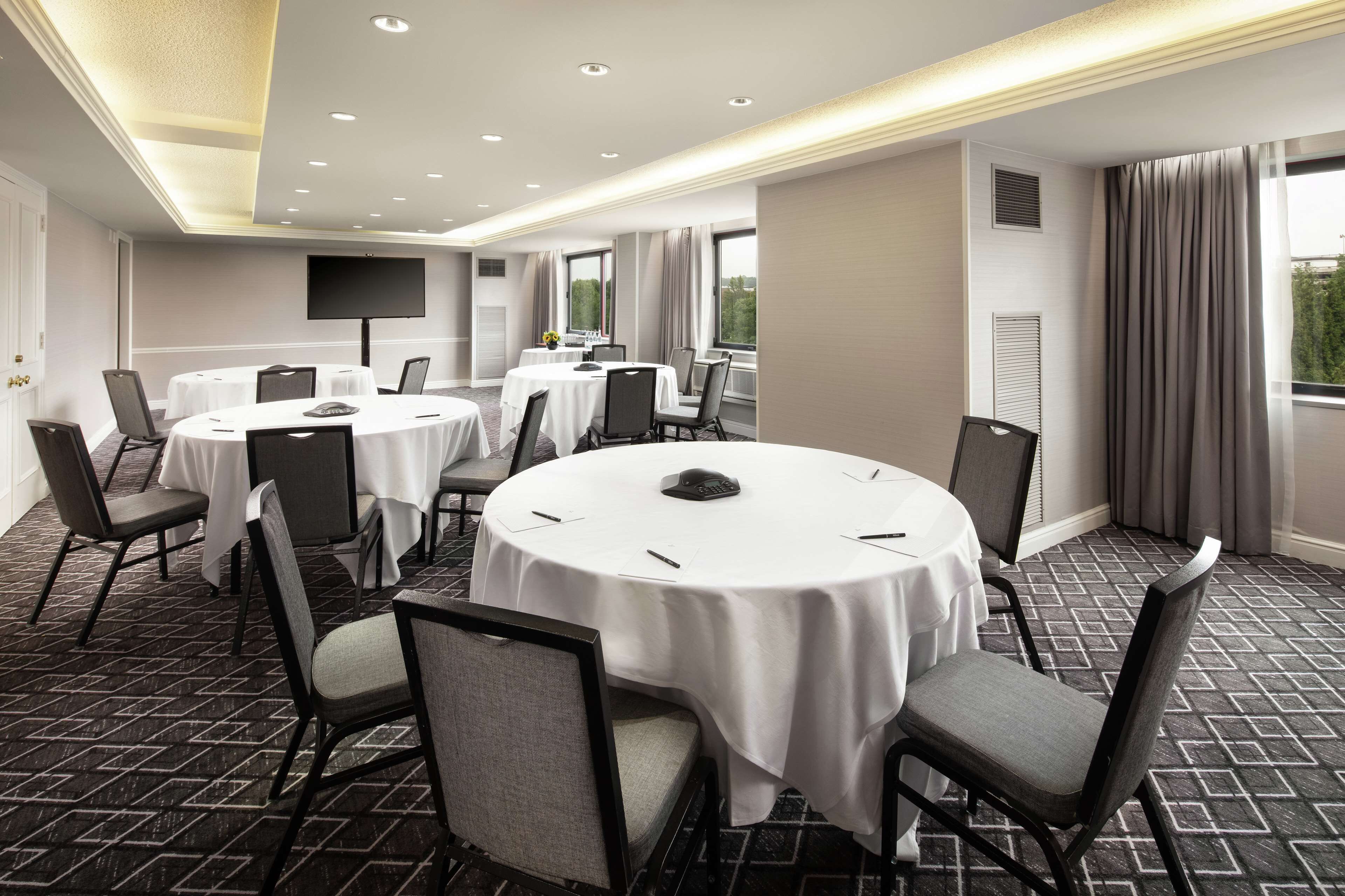 A conference room with round tables, chairs, a flat-screen TV, and large windows with curtains.
