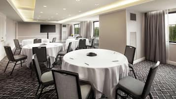 A conference room with round tables, chairs, a flat-screen TV, and large windows with curtains.