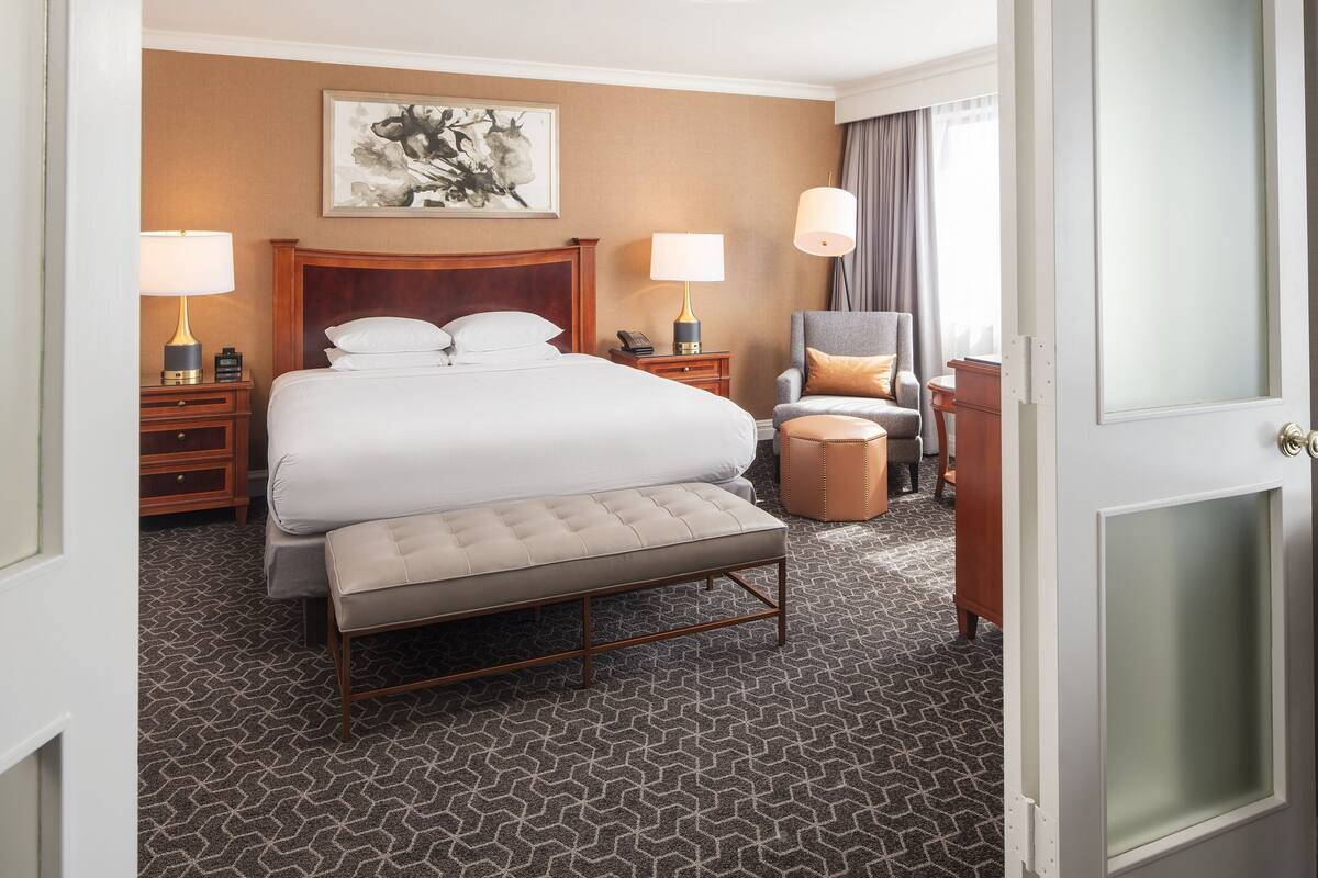 A hotel room with a large bed, two bedside tables with lamps, a chair, a bench, and a painting on the wall.
