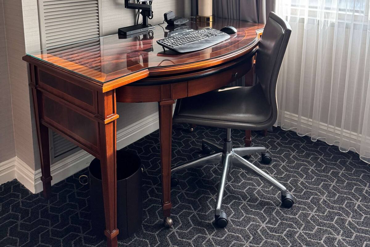 A hotel room with a wooden desk, a computer, a chair, a lamp, and a window with curtains.
