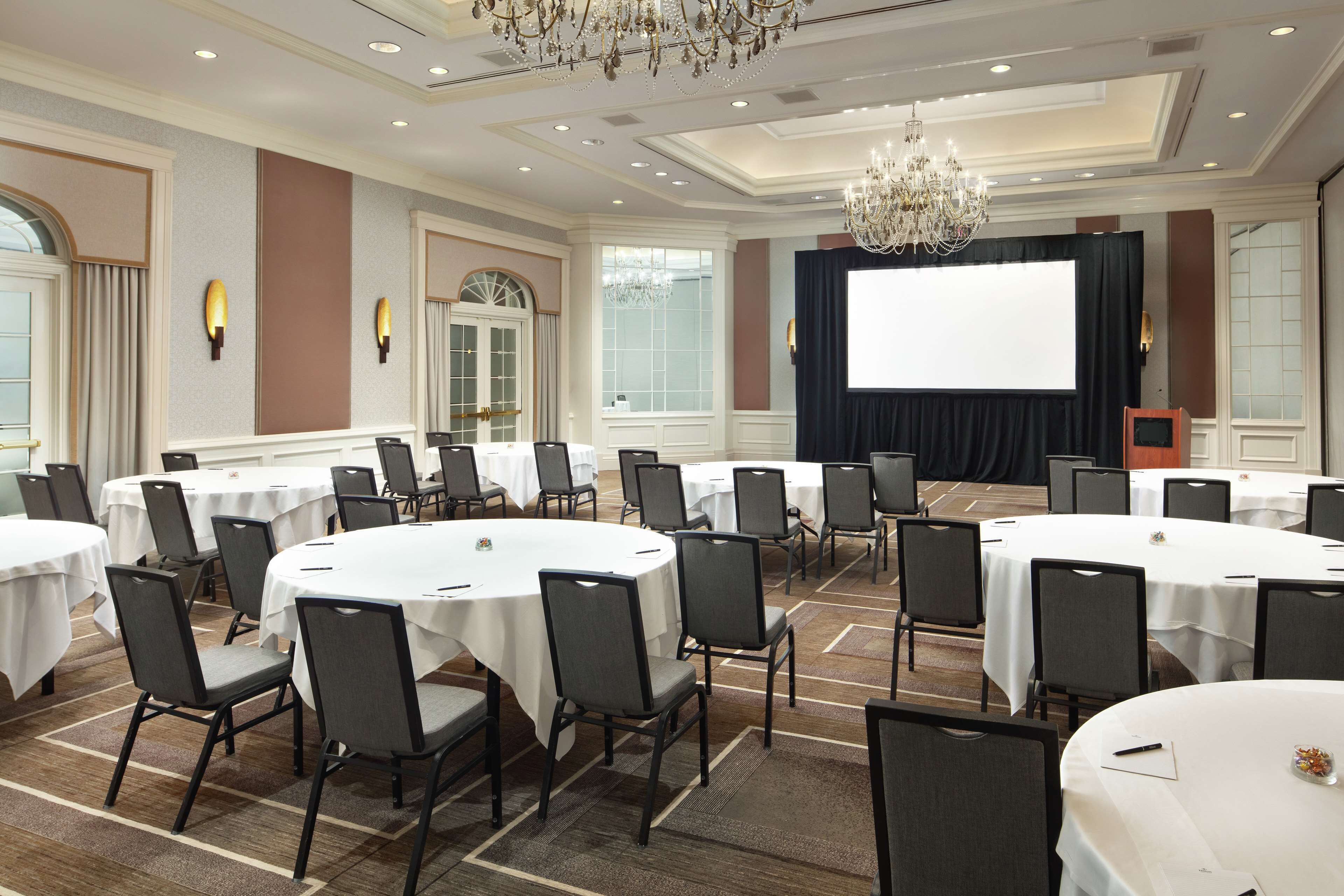 Un salón de conferencias con mesas redondas, sillas, una pantalla de proyector y un candelabro.