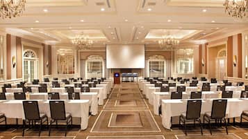 A large conference room with rows of tables and chairs, chandeliers, and a stage at the front.
